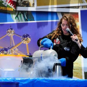 A curly haired woman in a black lab coat pours dry ice into a pot on a blue table