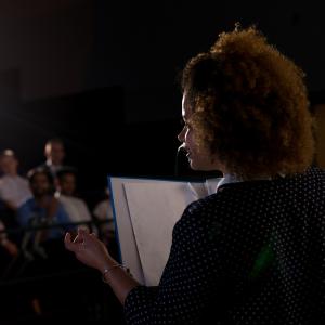 A woman presenting to a crowd in a darkened room while holding notes