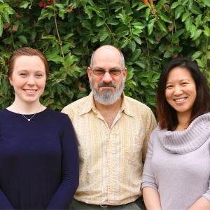 Two women and a man stand shoulder to shoulder in front of an ivy covered background. The man is standing in the middle. 