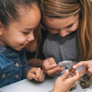 A close up of two girls using a magnifying glass to look at preserved bugs