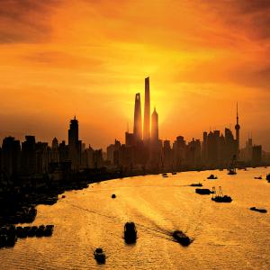 An orange filtered sunset image of a city on a river. The sun is setting directly behind three large spire sky scrapers