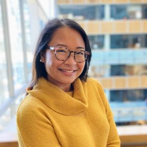 An asian person, who identifies as she/her they/them, smiles while wearing glasses and a yellow sweater. They are in a building setting with windows and multiple floor levels behind them. 