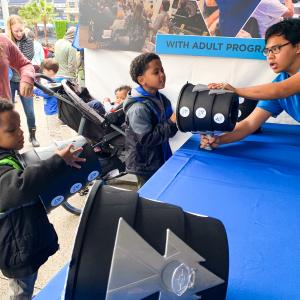 Two young children inspect a container with the assistence of a fleet science center educator at an outdoor community event.