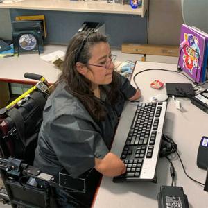 A dark haired woman sits at a desk with a computer keyboard. She is in a black wheelchair and is missing her forearms on both arms.  