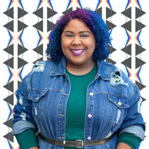 A black and hispanic woman with curly purple and blue hair stands facing the camera and smiles. The background behind her is a repeating pattern of triangles. 