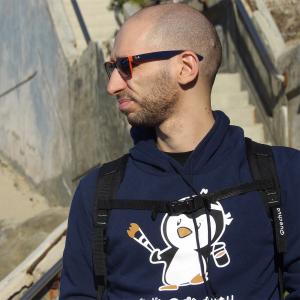 A bald white man stands in profile. He is wearing sunglasses and has light stubble. He is wearing a black backpack over a navy blue sweatshirt with a cartoon penguin holding a paintbrush on it. The background is an angle of concrete stairs leading up.