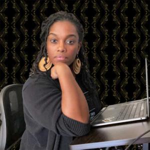 A black woman sits at a computer desk. He chin rests on her wrist and she faces the camera. She has long black hair and large gold earrings. There is a repeating black and yellow pattern behind her.