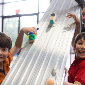 Two children and one adult race handmade vehicles down a metal slide.