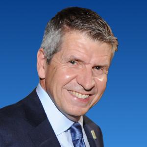 A middle aged white man smiles in a three quarter turn. He has short cropped salt and pepper hair. He is wearing a grey suit with a white shirt and a light blue tie. The background is a standard blue gradient profile background.  