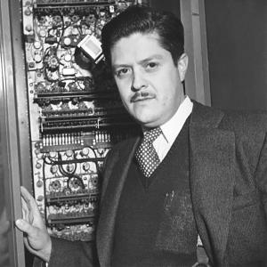 A black and white photo of a hispanic man with a mustache wearing a suit, sweater, and tie. He is standing in front of a wall of gears and mechanics.