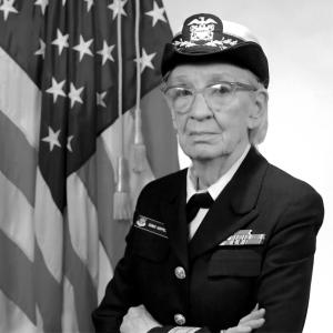 A black and white photo of an elderly white woman in a high ranking miltary uniform and hat. She is wearing glasses and has her arms closed. The American flag is hanging behind her.