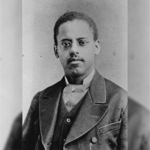 An old black and white photo of a black man with glasses and a mustache. He is wearing a jacket and suit. 