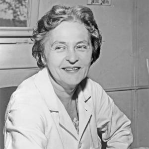 A black and white photo of a middle aged white woman looking to the left of the image while facing forward. She is wearing a white lab coat and is seated.