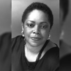 A black and white photo of a black woman. She is looking kindly at the viewer, has short cropped hair, round earrings and a multi strand necklace.