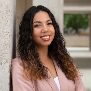A hispanic woman wearing a light pink blazer with dark curly hair leans against a concrete building an smiles.