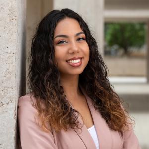 A hispanic woman wearing a light pink blazer with dark curly hair leans against a concrete building an smiles.  