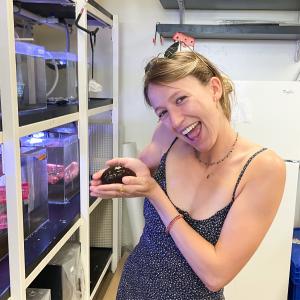 A white blonde woman holds a sea cucumber in her cupped hands and leans towards the camera with a large open mouthed smile. She has sunglasses on her head, two small necklaces, and is wearing a blue and white dotted summer dress. She is in a research style room with aquarium tanks on the left side and a white refrigerator behind her.