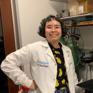 A woman in a white lab coat with short dark and green curly hair smiles at the camera. She is wearing a button up shirt that is black with bananas on it. The white lab coat has embroidered blue text that says Sanford Consortium for Regenerative Medicine UC San Diego. The background is a lab setting with large green canisters and shelves with boxes. 