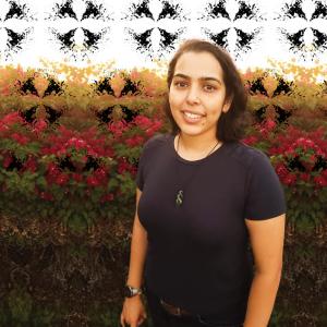 A tan skinned woman with shoulder length dark hair wearing a dark blue tshirt stands in front of a hedge row of green leaves and red flowers. A ink blot style pattern in black is behind her and in front of the hedge.