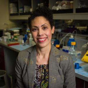 A hispanic woman with pinned up curly dark hair and pearl earrings smiles at the viewer. She is wearing a dark patterned blouse under a grey blazer with yellow embroidered flowers. She is sitting in a lab setting with bottles, tubes, and glassware behind her. 