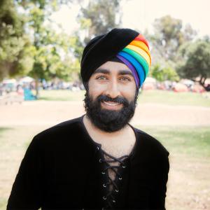 A Sihk man with a full dark beard and light eyes smiles at the camera. He is wearing a black long sleeved shirt with a string laced opening. His turban is black on the viewer left side and on the right, each fold is a separate color with red being towards the back then orange, yellow, green, blue, and finally purple closest to his forehead. He is in an outdoor park setting.  