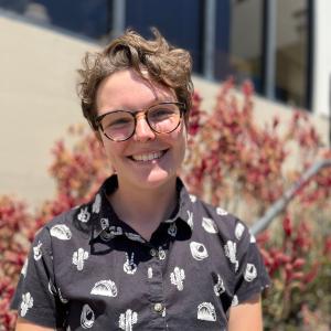 A person who's pronouns are She/Her and They/Them with short cropped brown wavy hair and glasses is wearing a black short sleeved button up shirt. The shirt has a pattern of white tacos, cactus, and avocados on it. They stand in front of the outside of a building where pink and red flowers are growing up the wall.  