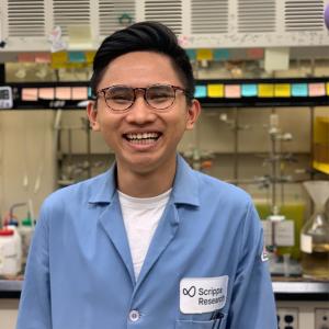 An asian man with glasses and short dark hair is smiling and wearing a blue lab coat. The lab coat has a white patch above the left breast pocket that says 'Scripps Research'. The man is standing in a lab setting. There are containers, jars, and vents in the background.