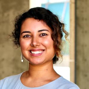 A Pakistani/Indian person whose pronouns are They/Them. They have curly dark hair that is pinned back behind their head. They are wearing a single silver earring on the viewer left that is a fish bone outline. They are wearing a light blue top and are standing outside near concrete walls. 