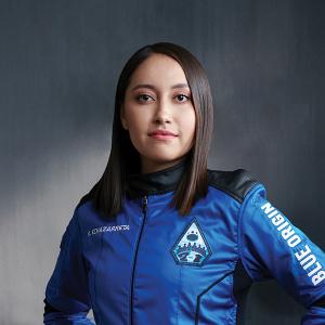 A young Mexican woman with shoulder length brown hair looks proudly at the camera. She is wearing a blue jumpsuit with 'Blue Origin' on the arm and a citizen astronaut patch on her left breast pocket. The background is a vague dark grey photography background.