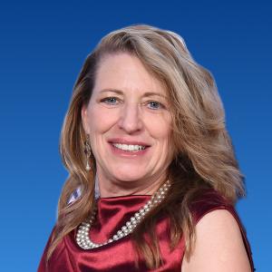 A white blond middle aged woman smiles. She is wearing a satin red top with a three rows of pearls necklace. The background is a default blue gradient with the darker blue on top. 
