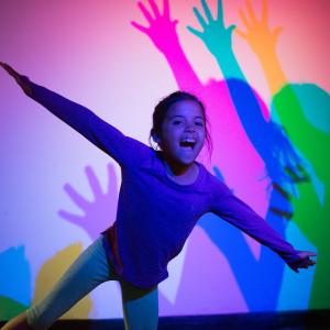 A young girl leans forward with her arms out and mouth open. He shadow behind her is in pink, blue, green, and yellow.