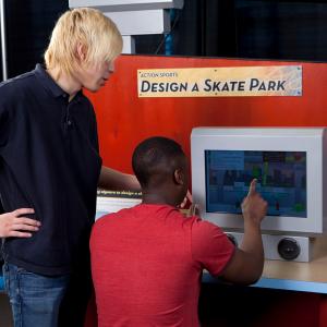 A white teen boy stands by a black teen boy. The black boy is sitting and touching a touch screen computer. Above them is a sign that says Design a Skate Park.