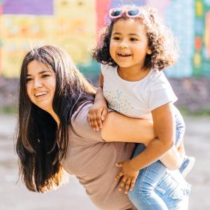 A hispanic woman with long dark hair smiles and holds a hispanic toddler girl on her back. They are outside with a blurry colorful mural behind them.