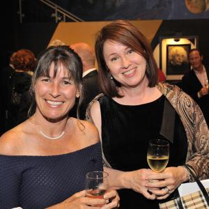 Two white women dress in cocktail attire lean together and smile at the viewer. They are each holding a glass of white wine.