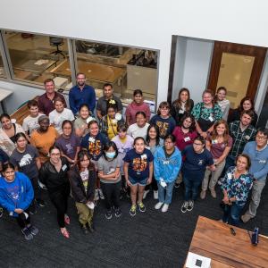 A large group of teen female students taken from an aerial view. There a few adults in the crowded group. They are in an office setting. Everyone is looking up at the camera and smiling.