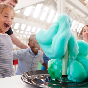 Four children looking at a chemistry science experiment in awe. The experiment is oozing blue and teal foam onto a table.