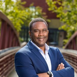 A middle aged black man in a blue business jacket and white collared shirt with arms crossed stands on a bridge and smiles at the camera