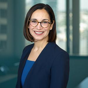 A young woman with short brown and glasses smiles at the camera. She is wearing a blue blazer over a blue shirt. 