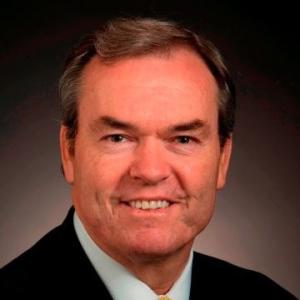 A middle aged white man in a black business jacket and white collared shirt smiles at the camera 