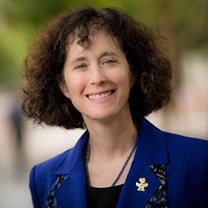 A white woman with chin length curly brown hair wearing an embroidered blue blazer and black top smiles at the camera 