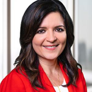 A white woman with dark brown hair wearing a red blazer and a white shirt smiles at the camera 