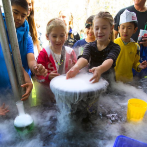 A group of eight students looking on and participating in a liquid nitrogen chemistry experiment in an outdoor setting. One student has their hands inside of a gray bowl overflowing with vapors.
