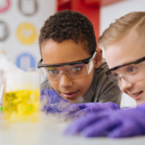 Two students focusing on a chemistry experiment inside of a lab. Both are wearing protective goggles and gloves.