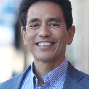 An asian man in a blue jacket and blue strip shirt smiles at the camera