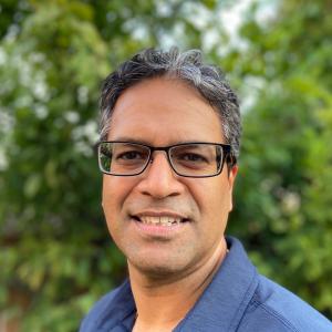 An indian man in a blue polo shirt with glasses and graying hair smiles at the camera outside