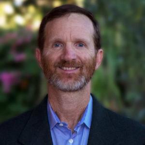 A middle aged white man with red hair and beard wearing business clothing smiles at the camera 