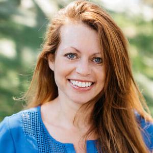 A white woman with long red hair and a blue shirt smiles at the camera 