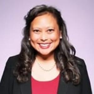 A woman with curled dark brown hair wearing a black blazer and red shirt with a pearl necklace smiles at the camera on a purple background. 