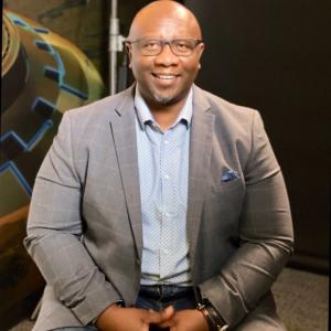 A middle aged black man wearing a grey jacket and blue collard shirt and glasses sits in front of a tech background and smiles at the camera 