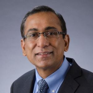 An indian man wearing glasses and a grey suit jacket with a blue collared shirt and blue tie smiles at the camera 